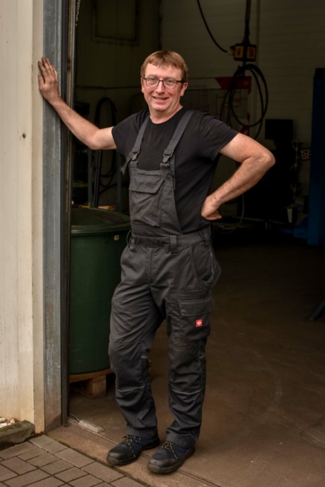 Dieter Motz steht lächelnd mit einem Arm an das Werkstatttor gelehnt.