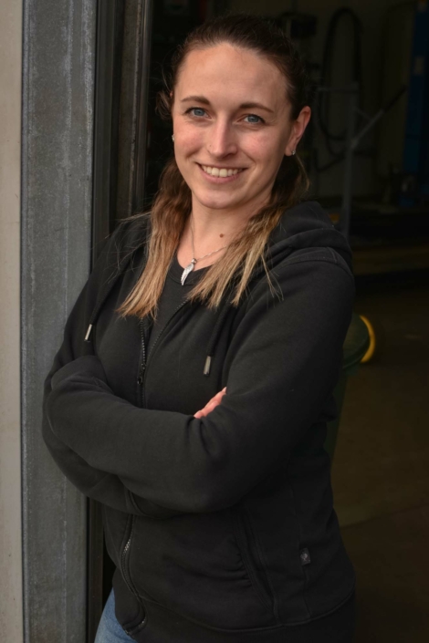 Julia Schott lehnt mit verschränkten Armen, lächelnd an der Innenseite des Werkstatttores.
