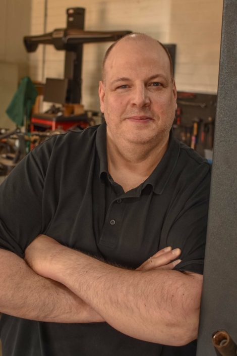 Mark Hoch lehnt mit verschränkten Armen, lächelnd an der Hebebühne in der Werkstatt.