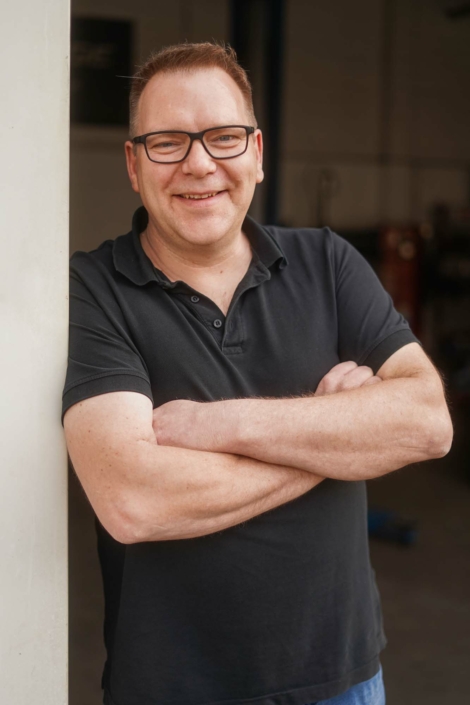 Rene Murk lehnt mit verschränkten Armen, lächelnd an der Innenseite des Werkstatttores.
